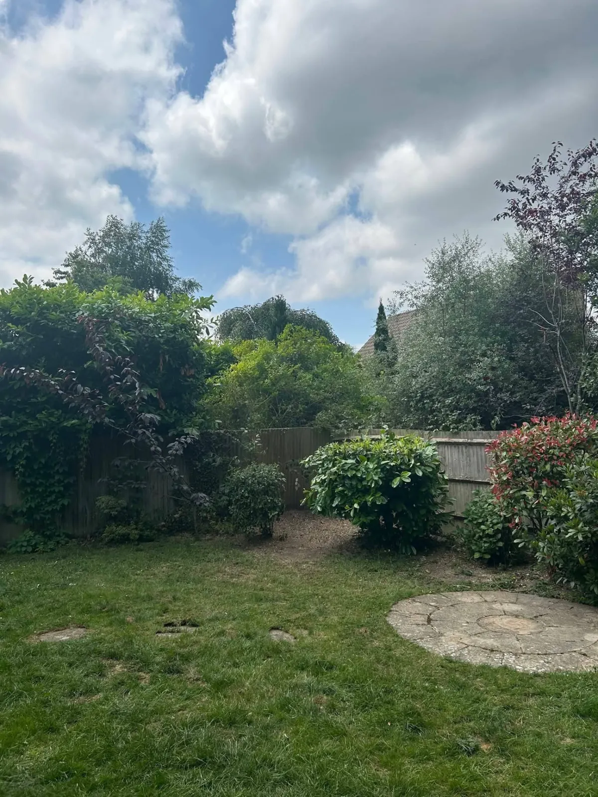 Open garden corner with lawn, shrubs and a circular paved feature under a wide sky